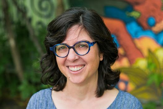Headshot of Stephanie Watt, cofounder and codirector, smiling with blue glasses and black shoulder-length hair. Wearing blue, a mural in background. 