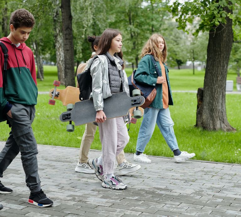 4 teenagers are walking on a footpath in a park with trees in the background. Two girls are carrying a skateboard. They are wearing backpacks. A diversity of teens in the photo.