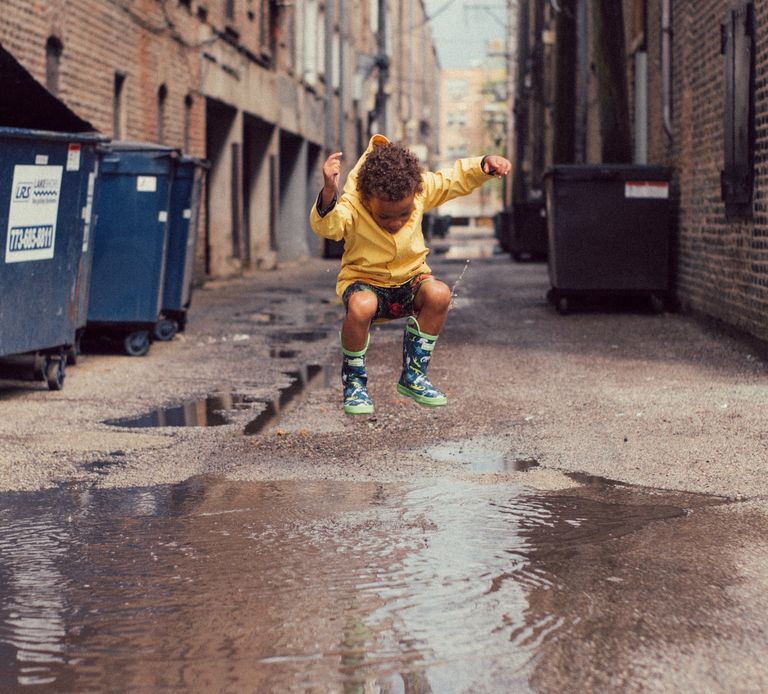 A brown child is jumping over a puddle in a commercial alleyway. The child is wearing a yellow jacket, blue and green rain boots and has arms up in the air.Big blue garbage bins are to the left..