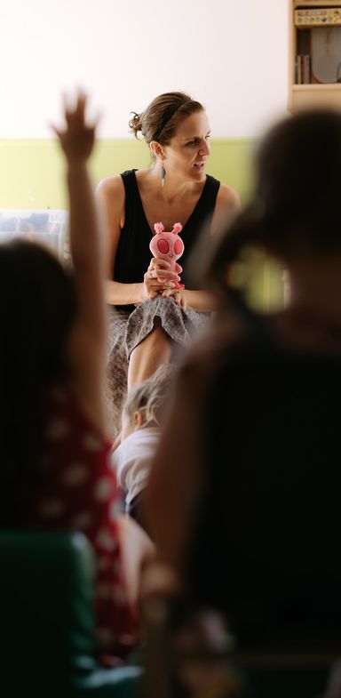 A white woman holding a teddy bear of an alien facilitating a public consultation among children. Children have their backs to the camera. One child has raised her arm to speak.