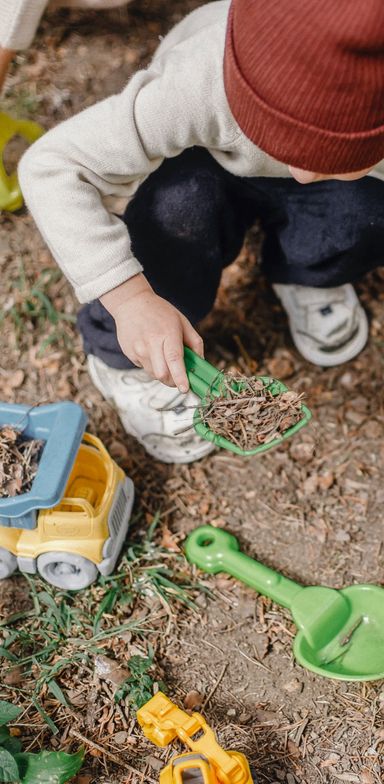 A child, seen from above, with a red beanie, playing in the dirt and wood chips with a small green shovel. Next to the child are two yellow trucks (plastic).