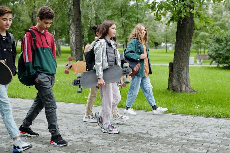 5 teenagers are walking on a footpath in a park with trees in the background. Two girls are carrying a skateboard. They are wearing backpacks. A diversity of teens in the photo.