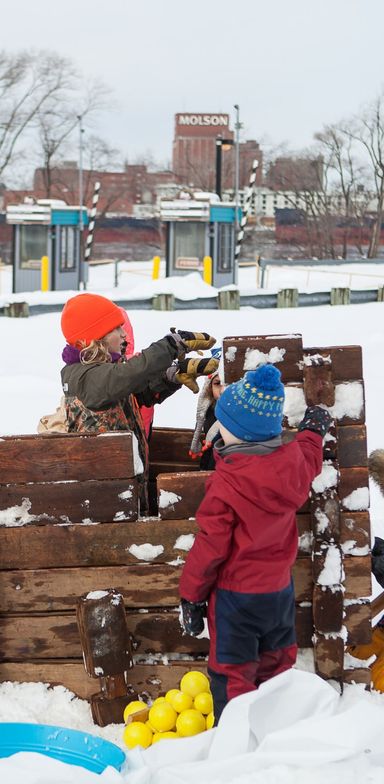 Deux enfants construisent une cabane avec des blocs de bois dans la neige. En arrière-scène, des guérites de stationnement et l'immeuble Molson à Montréal.