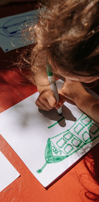 Enfant qui dessine une maison verte sur du papier blanc. On voit ses cheveux bruns de haut.