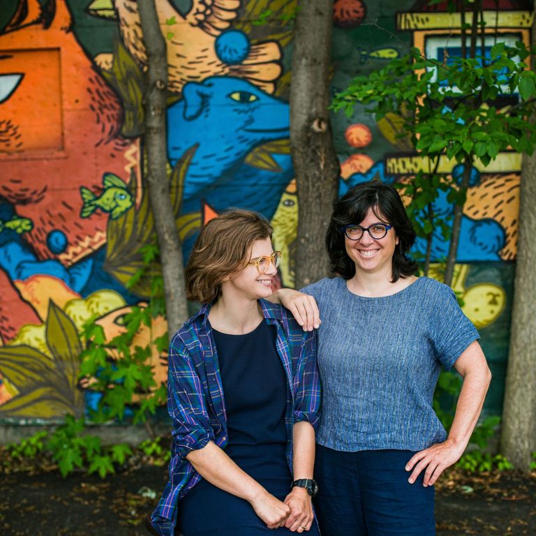 The two cofounders, Margaret Fraser sitting and looking to the right, Stephanie Watt standing and looking at the camera. They are both dressed in blue and are smiling. In the background is a mural of an orange fox and green vegetation.